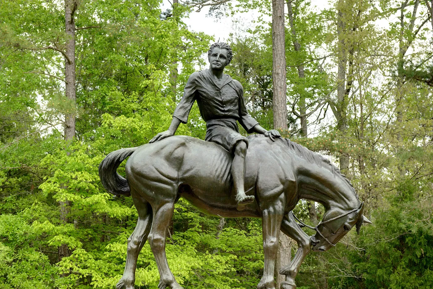 A bronze statue of a boy sitting on a grazing horse, set among bright green trees
