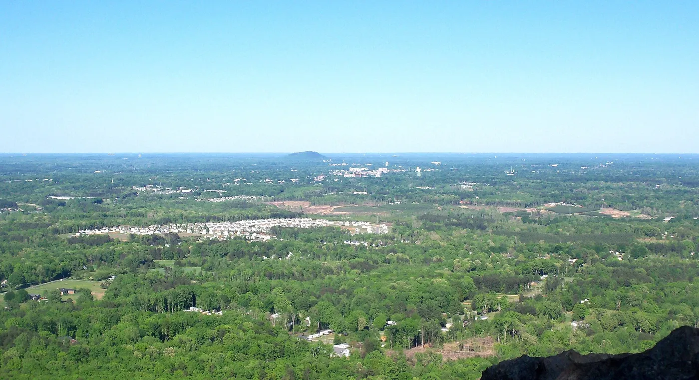 A wide view from a mountaintop over green Piedmont woods and suburbs, with a single distant hill on the horizon