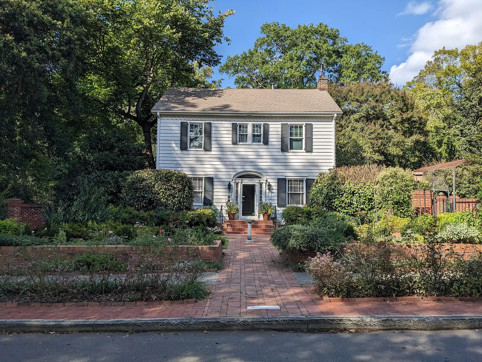A white two-story house with dark shutters behind a brick path and walled garden beds at Wing Haven